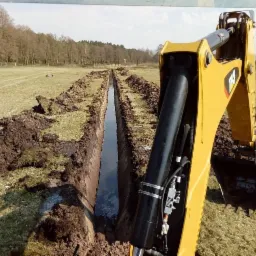 Żółta koparka Caterpillar z widocznym osprzętem hydraulicznym kopie wąski rów w zielonym terenie, tworząc kanał wypełniony wodą, z widocznym polem i drzewami w tle.