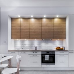 Usługi remontowe RJ - Nowoczesna kuchnia w Malborku z białymi szafkami dolnymi i drewnianymi górnymi, oświetlona punktowymi lampami podszafkowymi i kinkietem na ścianie, widoczny stół z nakryciem.