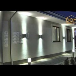 Usługi remontowe RJ - Elewacja domu z nowoczesnym oświetleniem zewnętrznym: kinkiety ścienne emitujące strumienie światła w górę i w dół, dodatkowe lampy stojące wzdłuż ścieżki. Tablica informacyjna widoczna na ścianie.