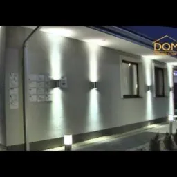 Elewacja domu z nowoczesnym oświetleniem zewnętrznym: kinkiety ścienne emitujące strumienie światła w górę i w dół, dodatkowe lampy stojące wzdłuż ścieżki. Tablica informacyjna widoczna na ścianie.