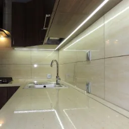 Nowoczesna kuchnia z beżowymi płytkami imitującymi marmur, podświetleniem LED pod szafkami i nad blatem, chromowaną baterią i gniazdkami elektrycznymi wkomponowanymi w płytki.