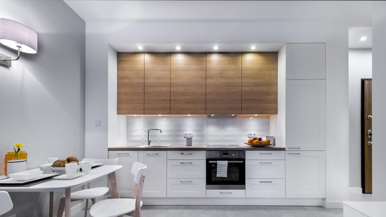 Nowoczesna kuchnia w Malborku z białymi szafkami dolnymi i drewnianymi górnymi, oświetlona punktowymi lampami podszafkowymi i kinkietem na ścianie, widoczny stół z nakryciem.