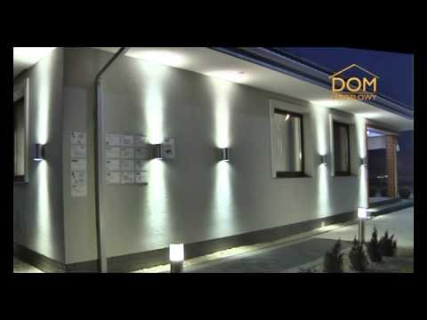 Elewacja domu z nowoczesnym oświetleniem zewnętrznym: kinkiety ścienne emitujące strumienie światła w górę i w dół, dodatkowe lampy stojące wzdłuż ścieżki. Tablica informacyjna widoczna na ścianie.