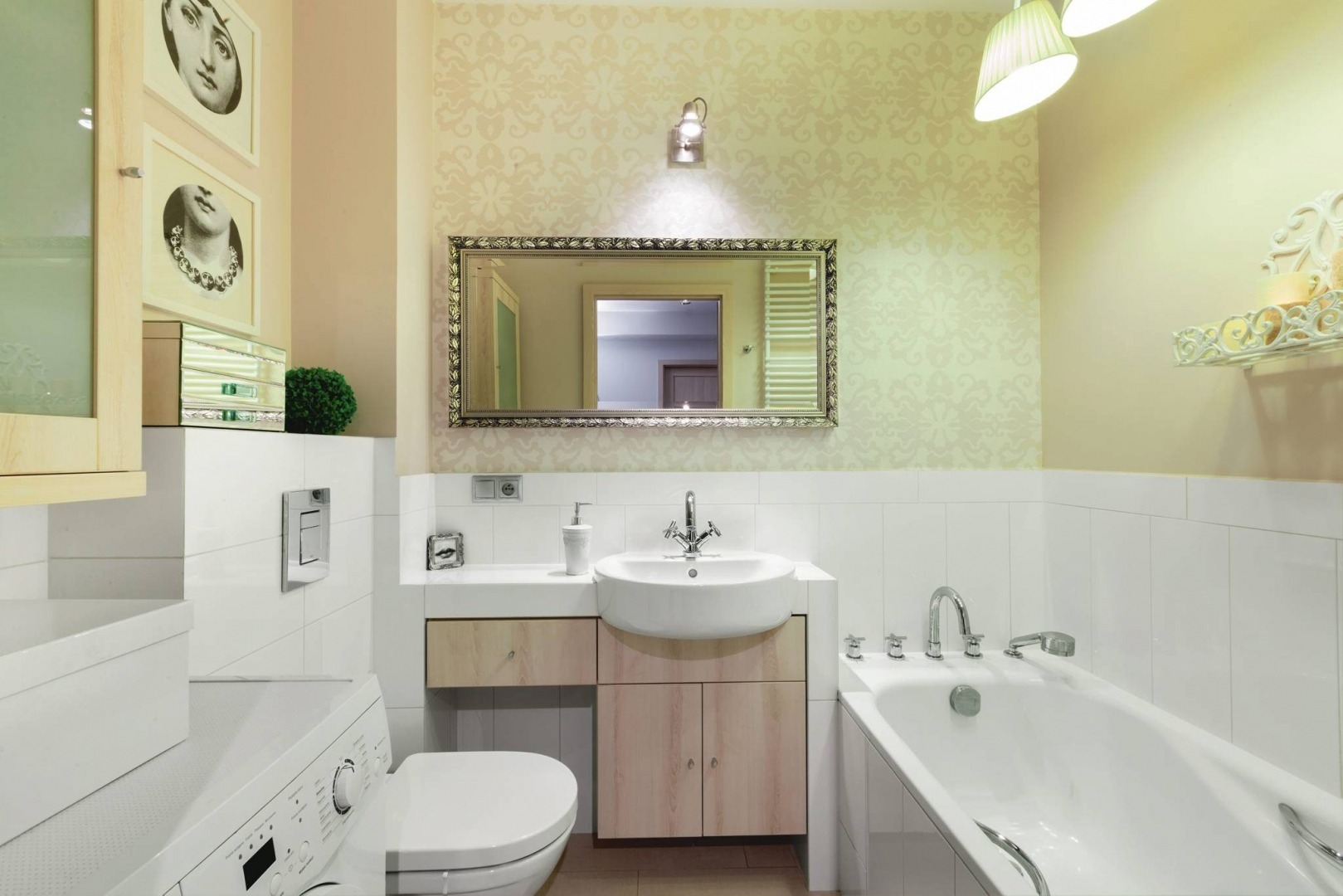 Jasna łazienka z wanną, umywalką w zabudowie z drewnianymi szafkami, toaletą i pralką; lustro w ozdobnej ramie i dekoracyjna tapeta w tle.