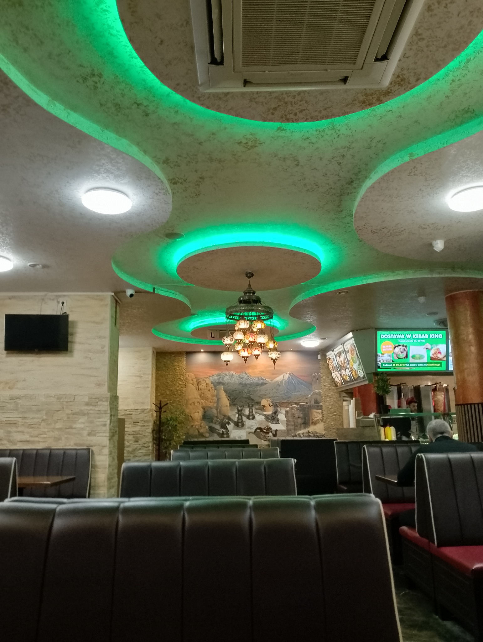 Wnętrze lokalu gastronomicznego z dekoracyjnym sufitem podwieszanym z zielonym oświetleniem LED, wiszącą lampą w stylu orientalnym i muralem na ścianie.