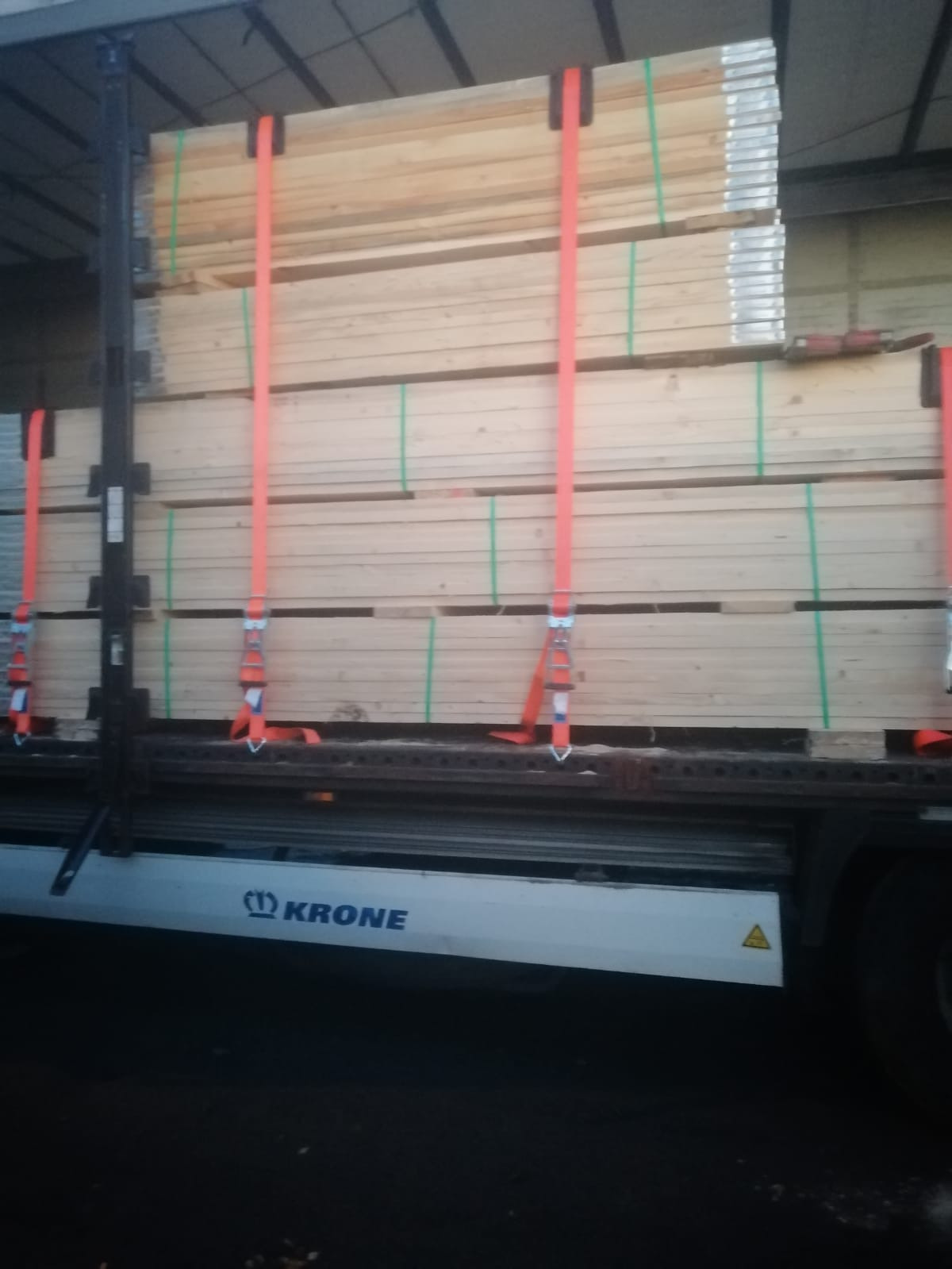 Załadunek desek na paletach zabezpieczonych pomarańczowymi pasami transportowymi na naczepie ciężarówki marki Krone.
