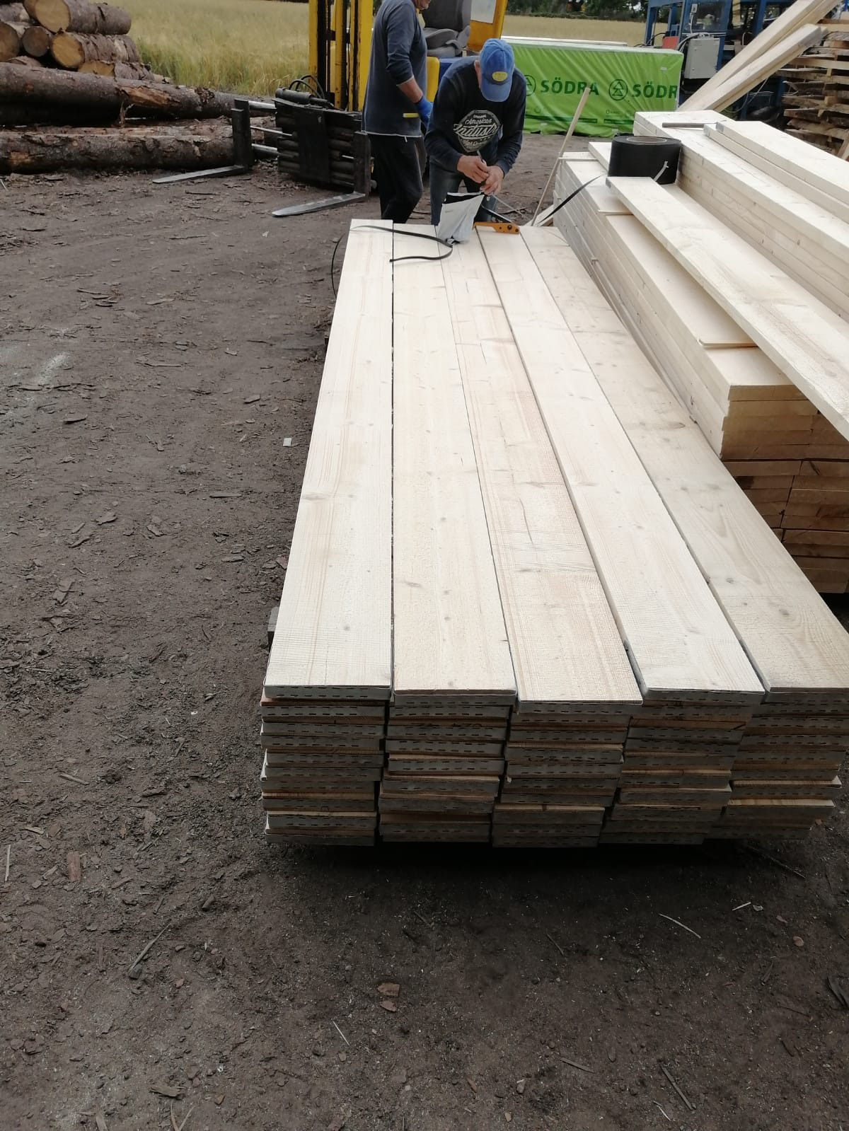 Stos świeżych, jasnych desek z widocznymi otworami montażowymi, przygotowywanych do załadunku przez dwóch pracowników w odzieży roboczej, z wózkiem widłowym w tle.