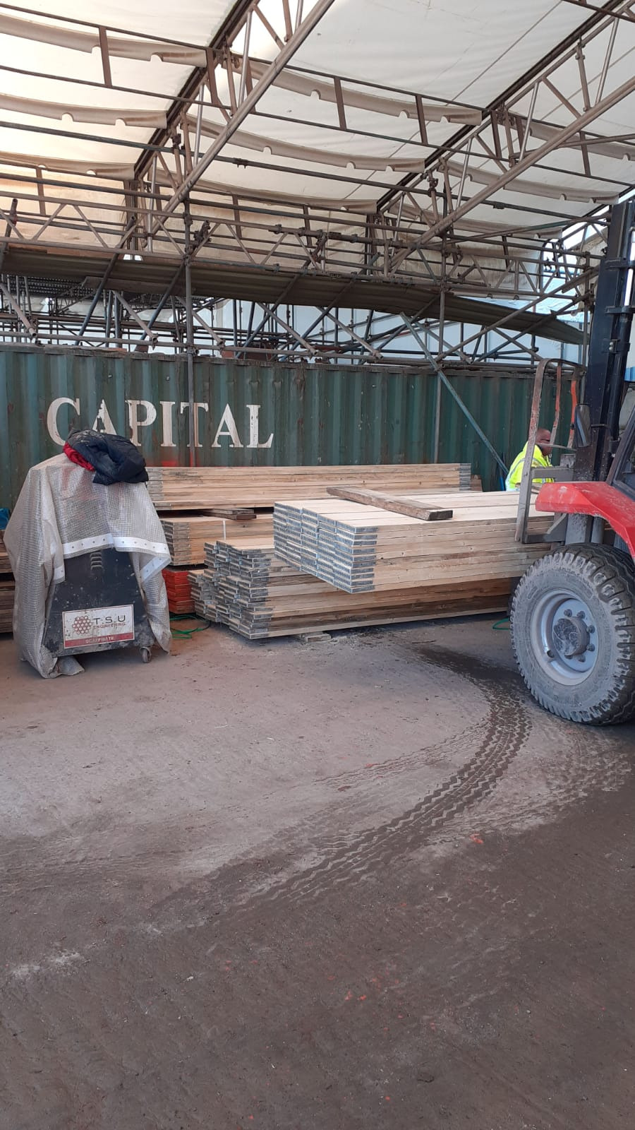 Składowane deski budowlane pod zadaszeniem, obok wózek widłowy i kontener z napisem CAPITAL, w tle rusztowania.