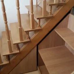 Drewniane schody z balustradą i toczonymi tralkami, zabudowane szafkami z półkami, widok z dołu.