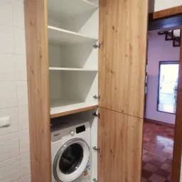 Szafa na wymiar z drewnianymi frontami, kryjąca pralkę i posiadająca białe półki, otwarte drzwi ukazują wnętrze i fragment schodów w tle.