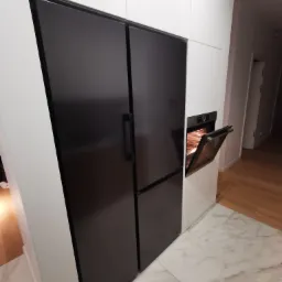 Nowoczesna zabudowa kuchenna z czarnym, dużym lodówko-zamrażarką side-by-side i piekarnikiem z otwartymi drzwiczkami, wbudowanym w białe szafki, na marmurowej podłodze.