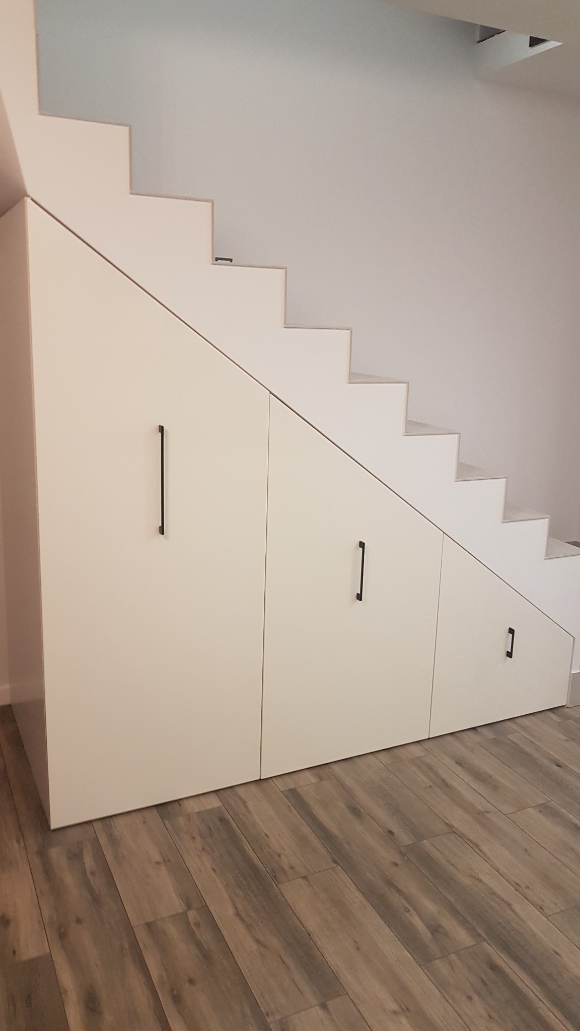 Białe szafki z czarnymi uchwytami, wbudowane pod schodami, na podłodze panele imitujące drewno.