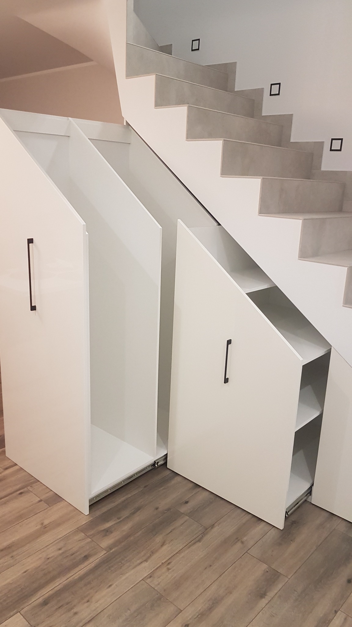 Białe, wysuwane szafki pod schodami z czarnymi, minimalistycznymi uchwytami, widoczne półki w jednej z otwartych szafek, na podłodze panele imitujące drewno.