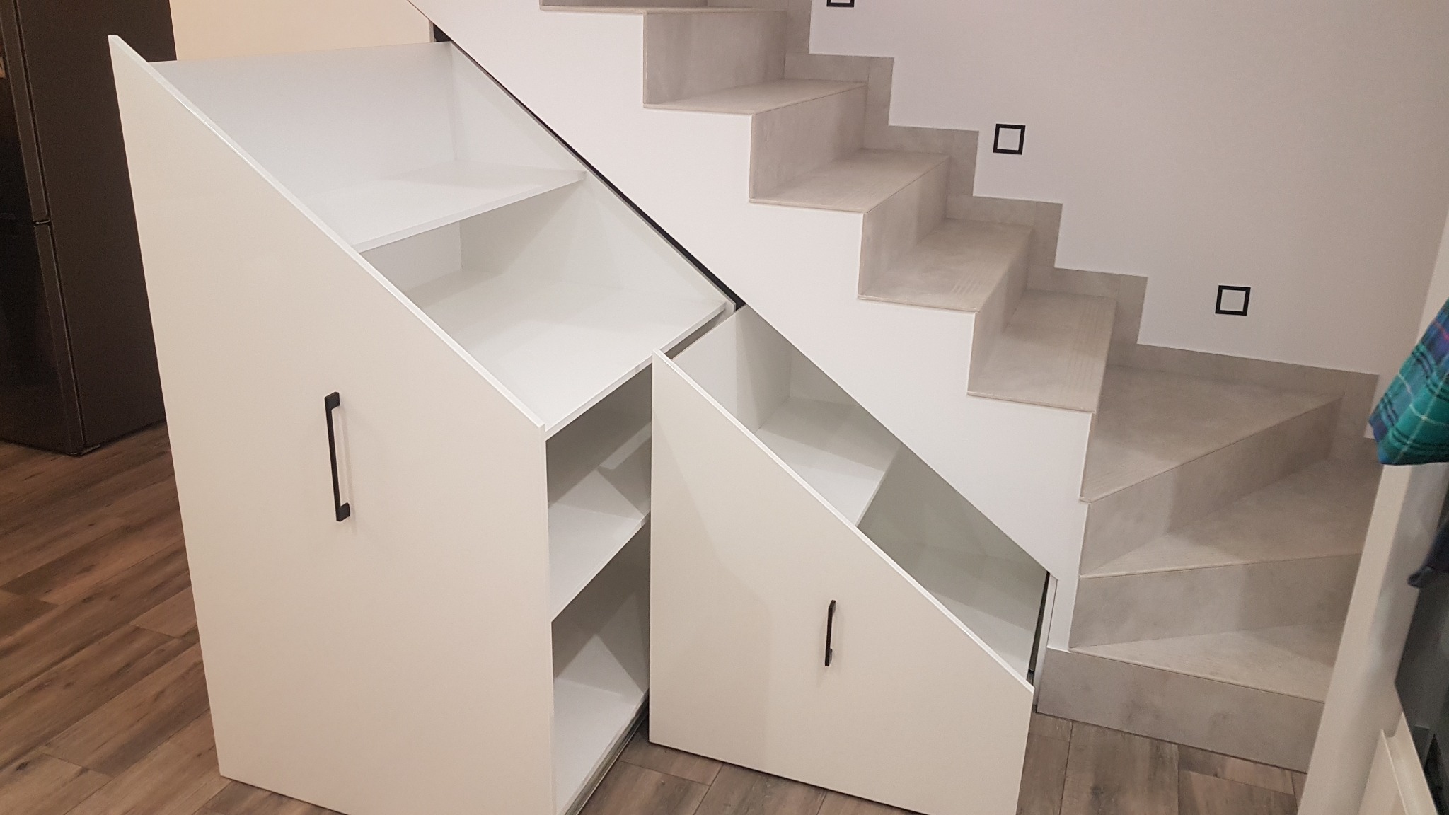 Białe szafki z półkami i szufladami wbudowane pod schody, z czarnymi, minimalistycznymi uchwytami.