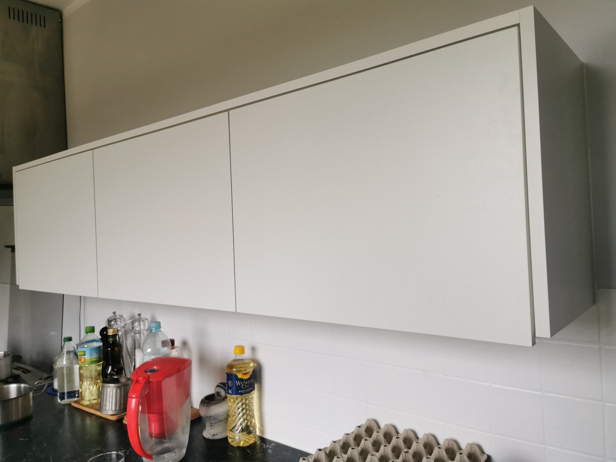 Białe szafki kuchenne wiszące nad blatem z akcesoriami kuchennymi, widoczne butelki z olejem, dzbanek filtrujący i pojemnik na jajka.