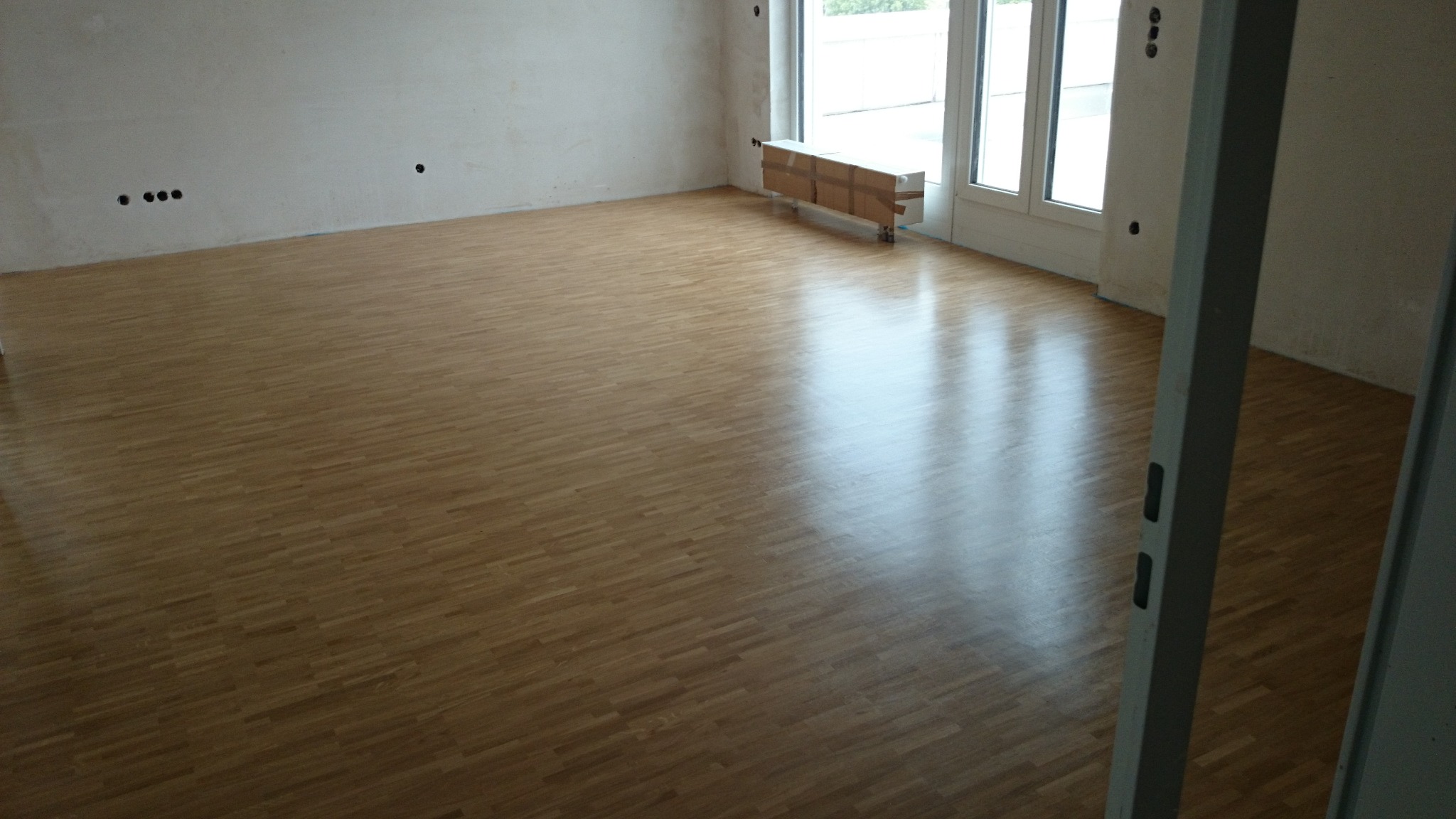 Świeżo położona, jasna podłoga z parkietu w pustym pokoju, z widocznymi śladami po niedokończonych pracach wykończeniowych na ścianach i osłoniętym grzejniku przy oknie.