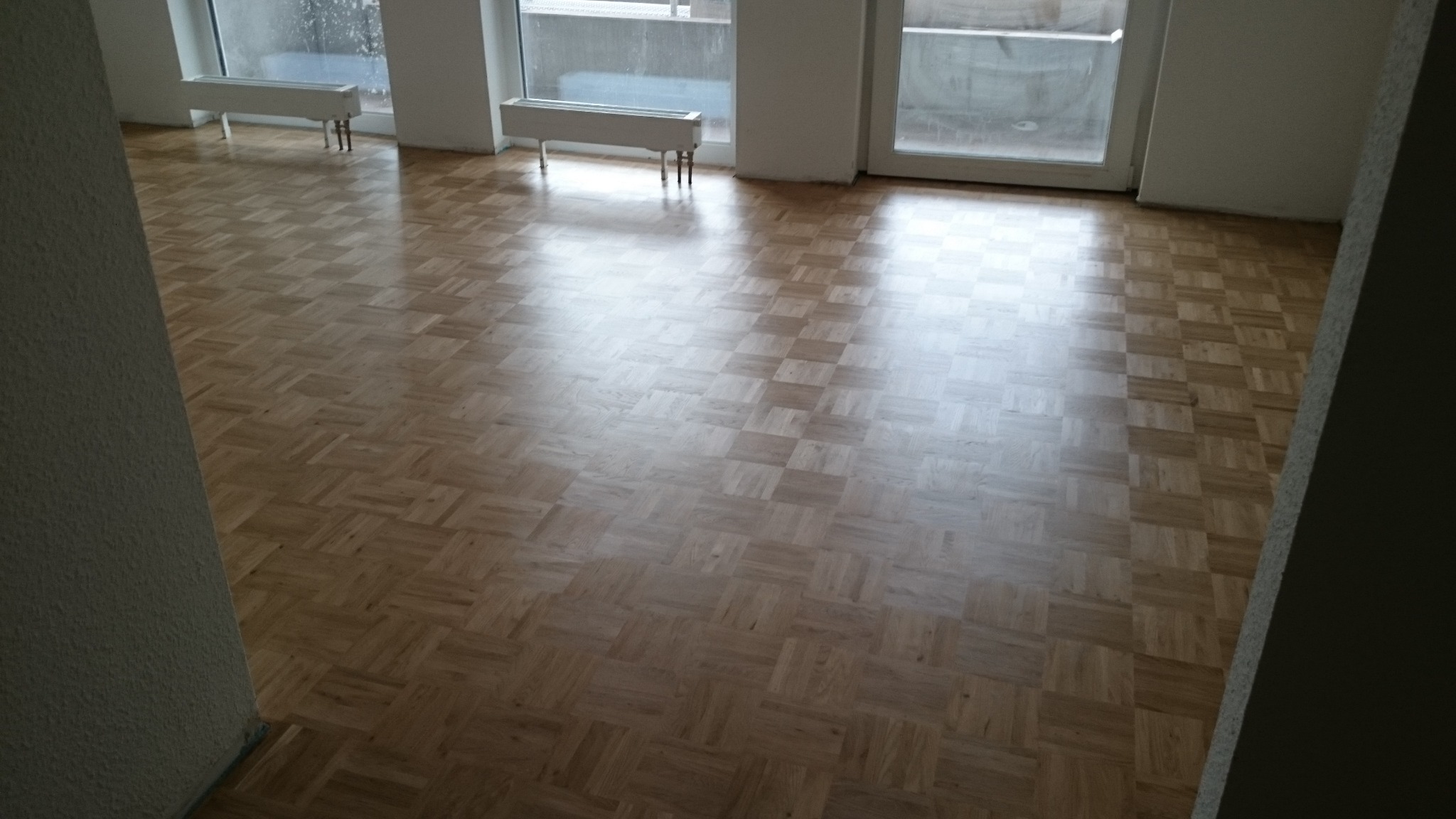 Parkiet ułożony w jodełkę w pustym pokoju z grzejnikami pod oknami.