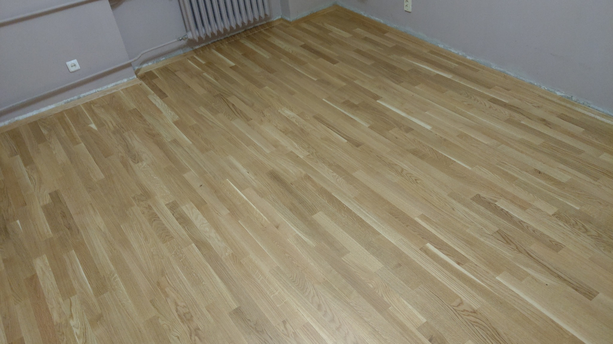 Jasny, drewniany parkiet ułożony w pokoju, widoczny grzejnik i gniazdko elektryczne na ścianie.