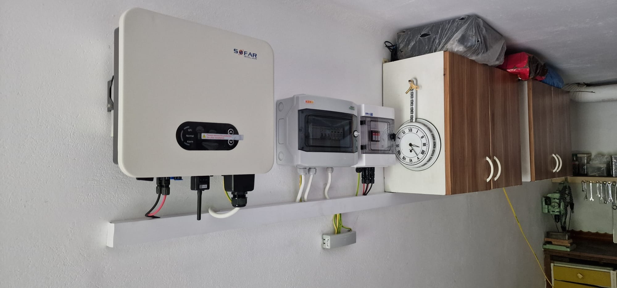 Biały inwerter SOFAR SOLAR zamontowany na ścianie obok szarych skrzynek elektrycznych i dekoracyjnego zegara ściennego, widoczne kable i przewody.