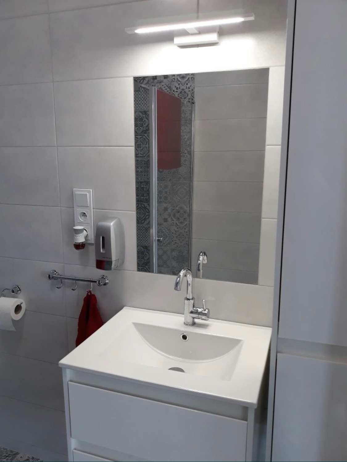 Biała umywalka z szafką w łazience po remoncie, bateria chromowana, widoczne płytki w odcieniach szarości i dozownik mydła.