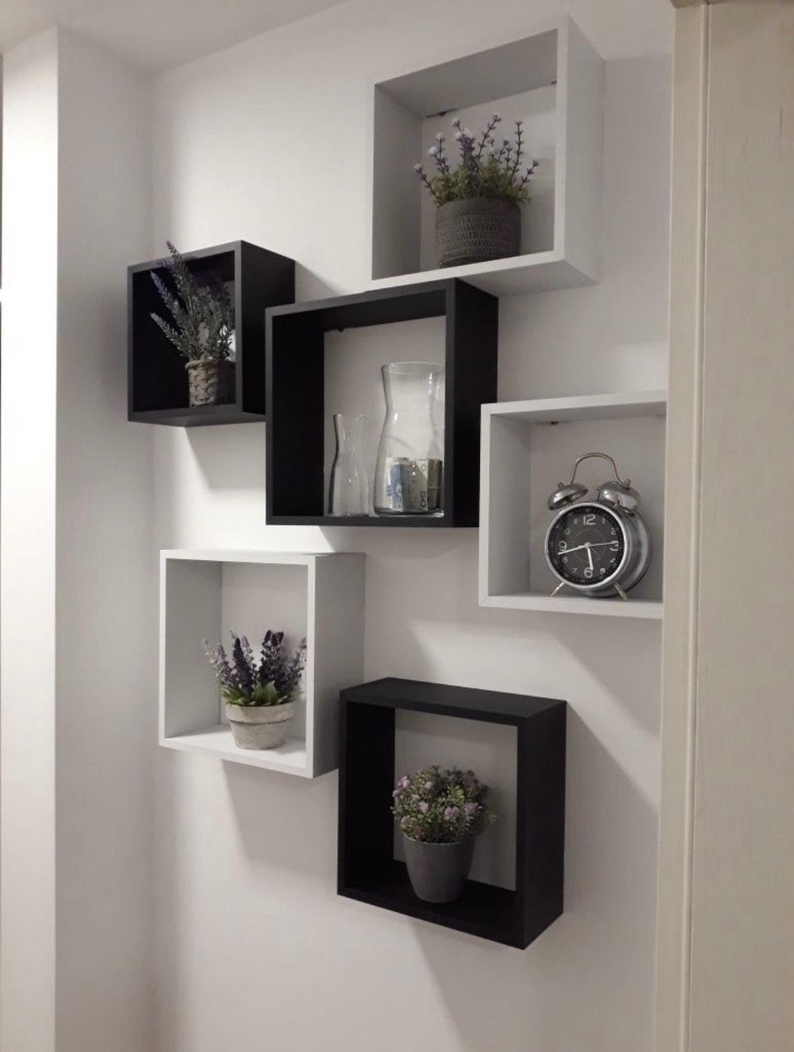 Aranżacja ściany z sześcioma kwadratowymi półkami w kolorach czarnym i białym, na których znajdują się dekoracje: kwiaty w doniczkach, szklane wazony i srebrny budzik.