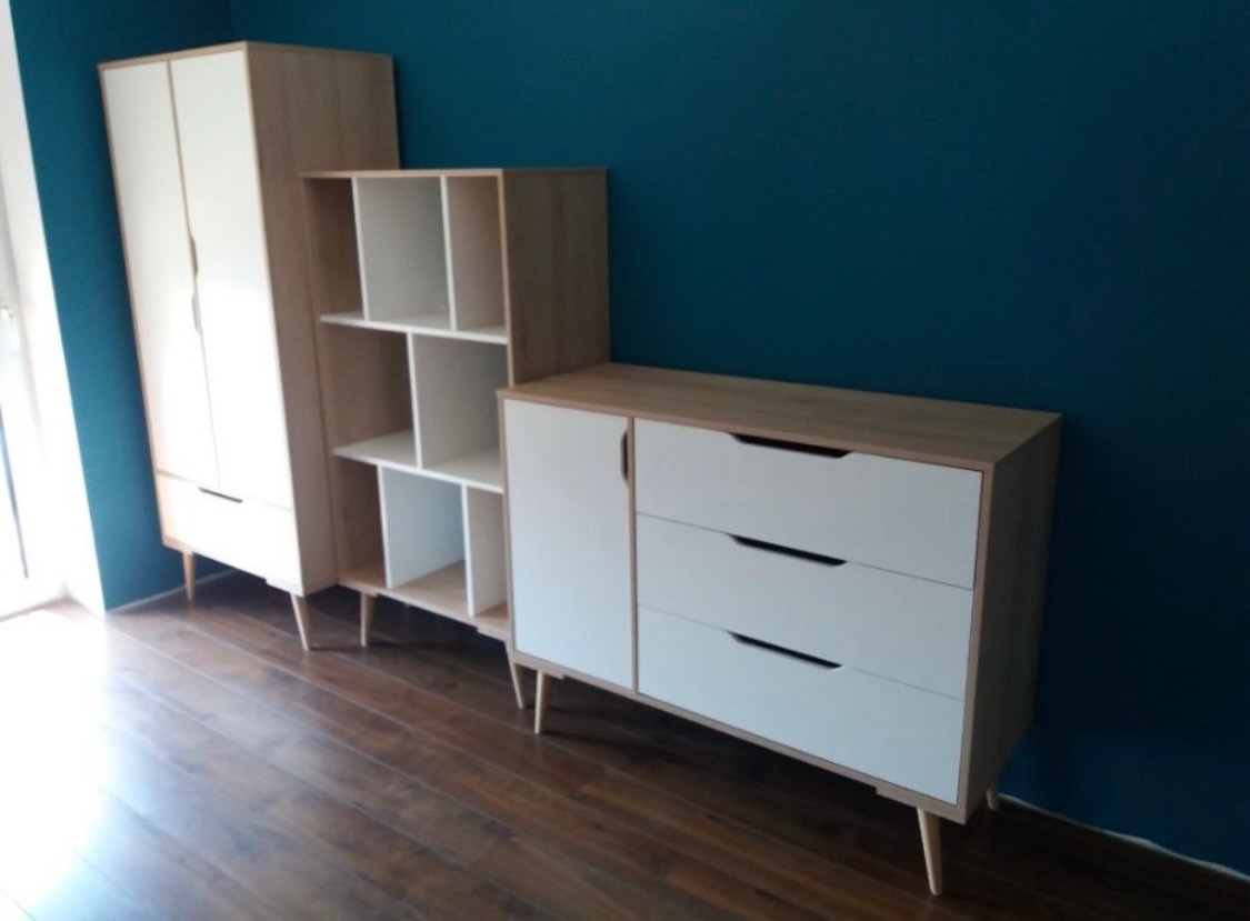 Ustawienie mebli dziecięcych: szafa, regał i komoda z białymi frontami i drewnianymi korpusami na tle ciemnoturkusowej ściany i drewnianej podłogi.