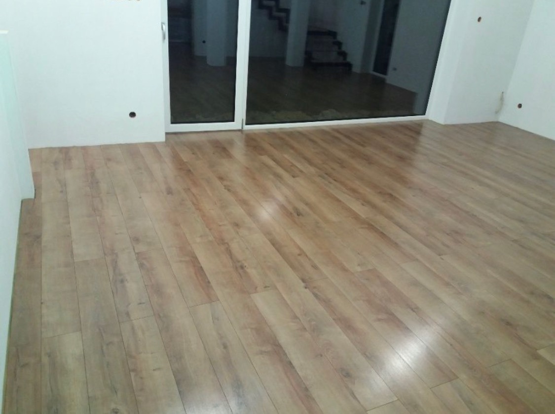 Nowo ułożona, jasna podłoga z paneli laminowanych w pustym pokoju z dużym oknem przesuwnym i widocznymi schodami w tle.