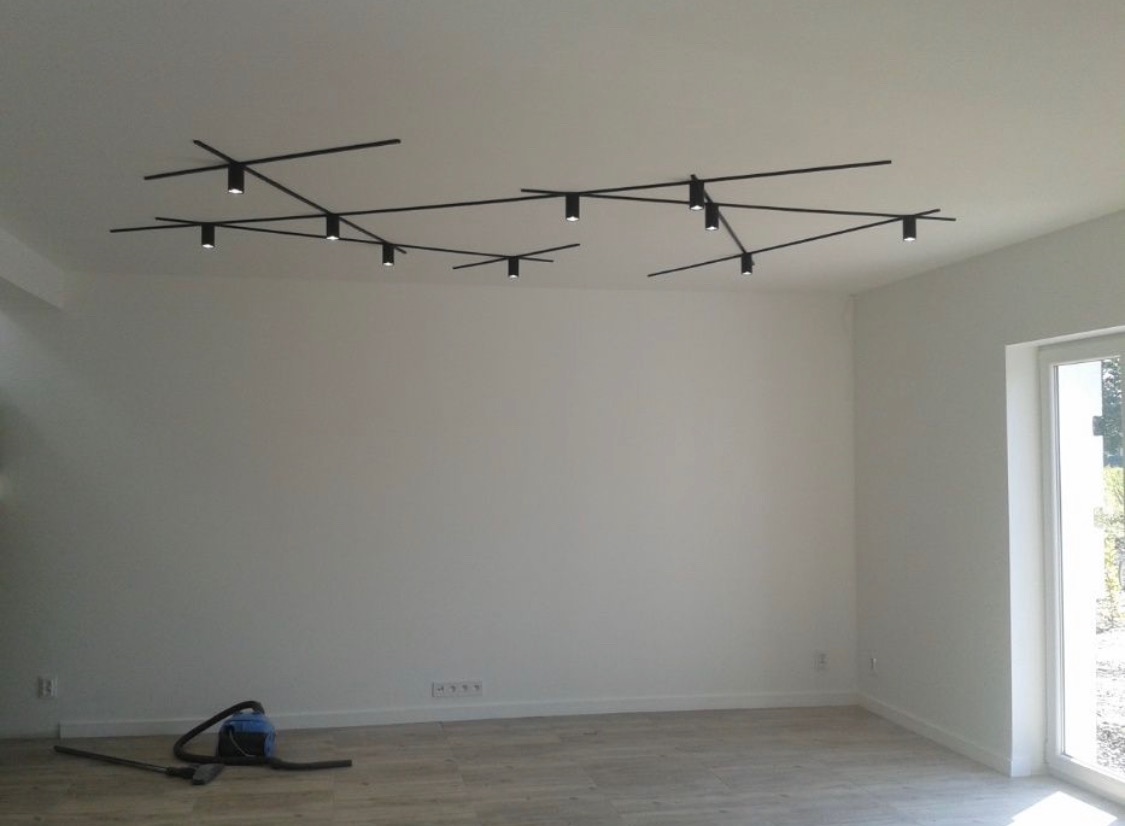 Minimalistyczne wnętrze z zamontowanym, nowoczesnym systemem oświetlenia szynowego w kolorze czarnym, z widocznym odkurzaczem na podłodze.