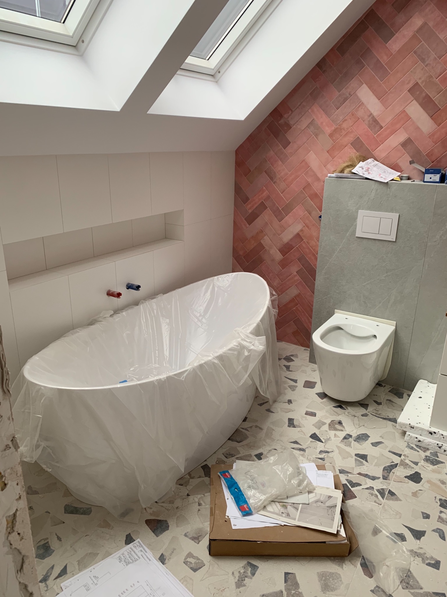 Łazienka w trakcie remontu: wolnostojąca wanna zabezpieczona folią, podwieszana toaleta, ściana z cegiełek ułożonych w jodełkę, mozaika na podłodze, plany i narzędzia na podłodze, okno dachowe.