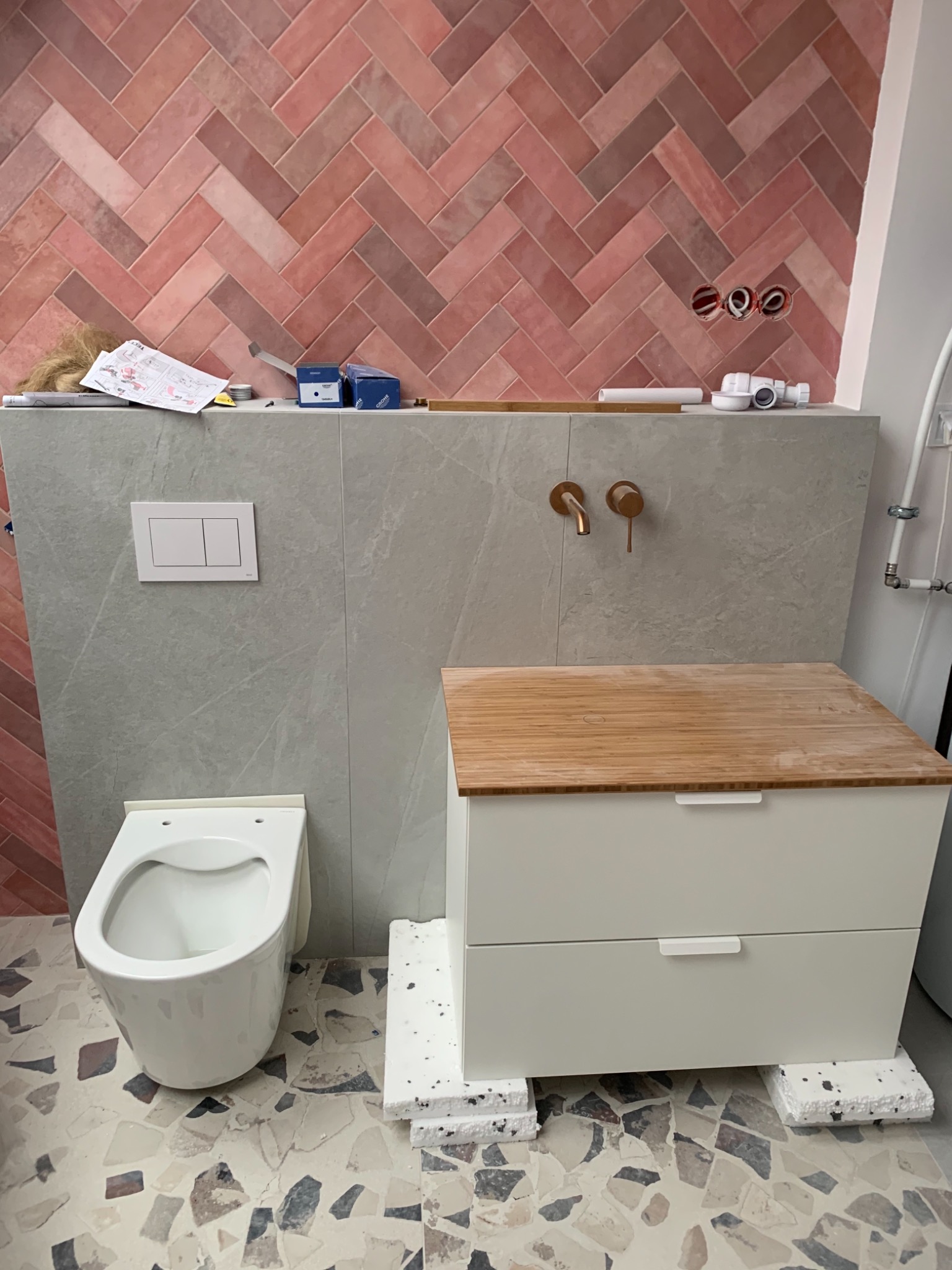 Niedokończona łazienka z różową cegłą ułożoną w jodełkę, wisząca toaleta, szafka pod umywalkę z bambusowym blatem, miedziana bateria i podłoga w stylu lastryko, widoczne elementy instalacji.