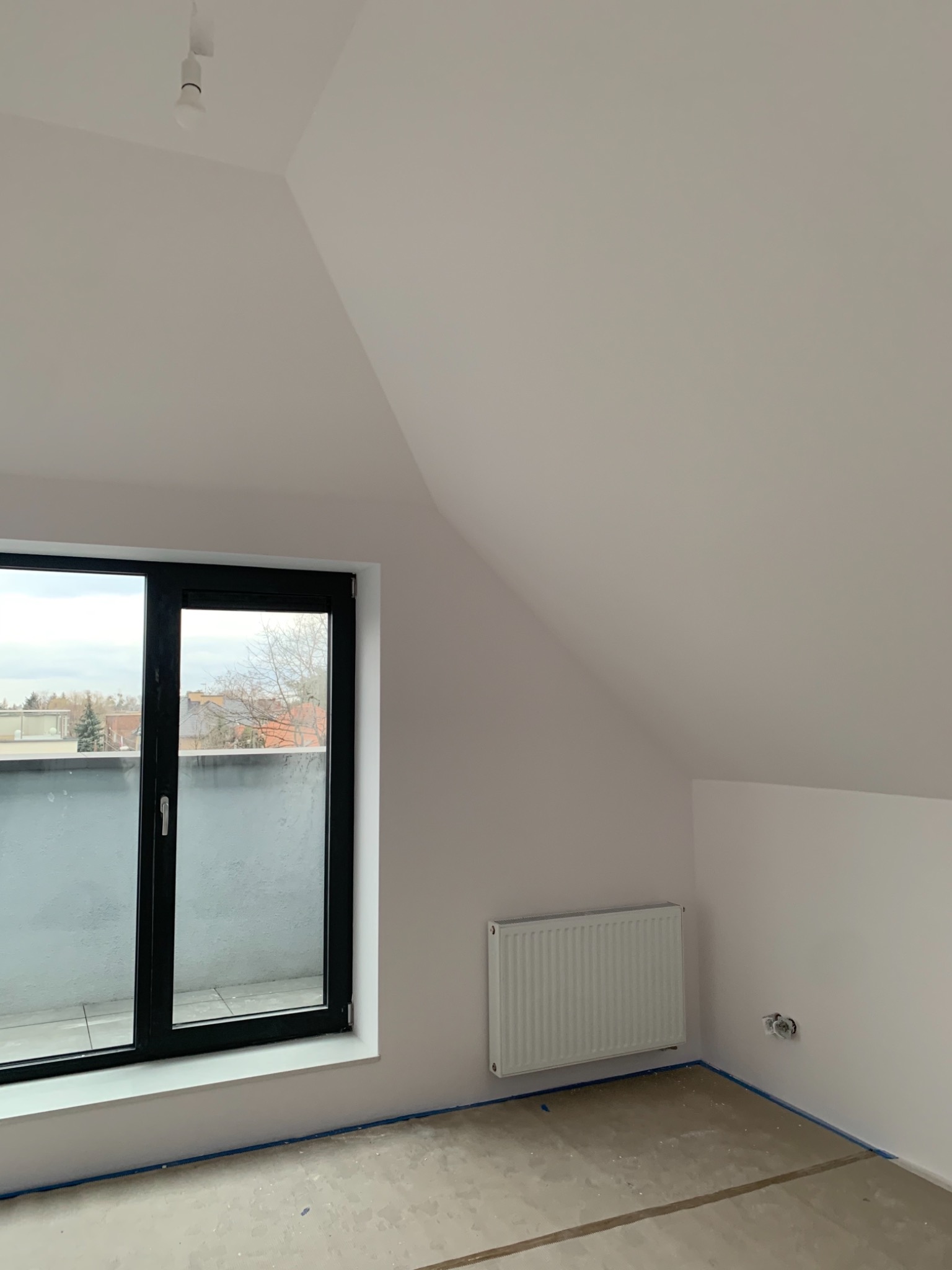 Poddasze po malowaniu na biało, widoczne okno balkonowe z czarną ramą, grzejnik i zabezpieczona folią podłoga przed dalszymi pracami wykończeniowymi.