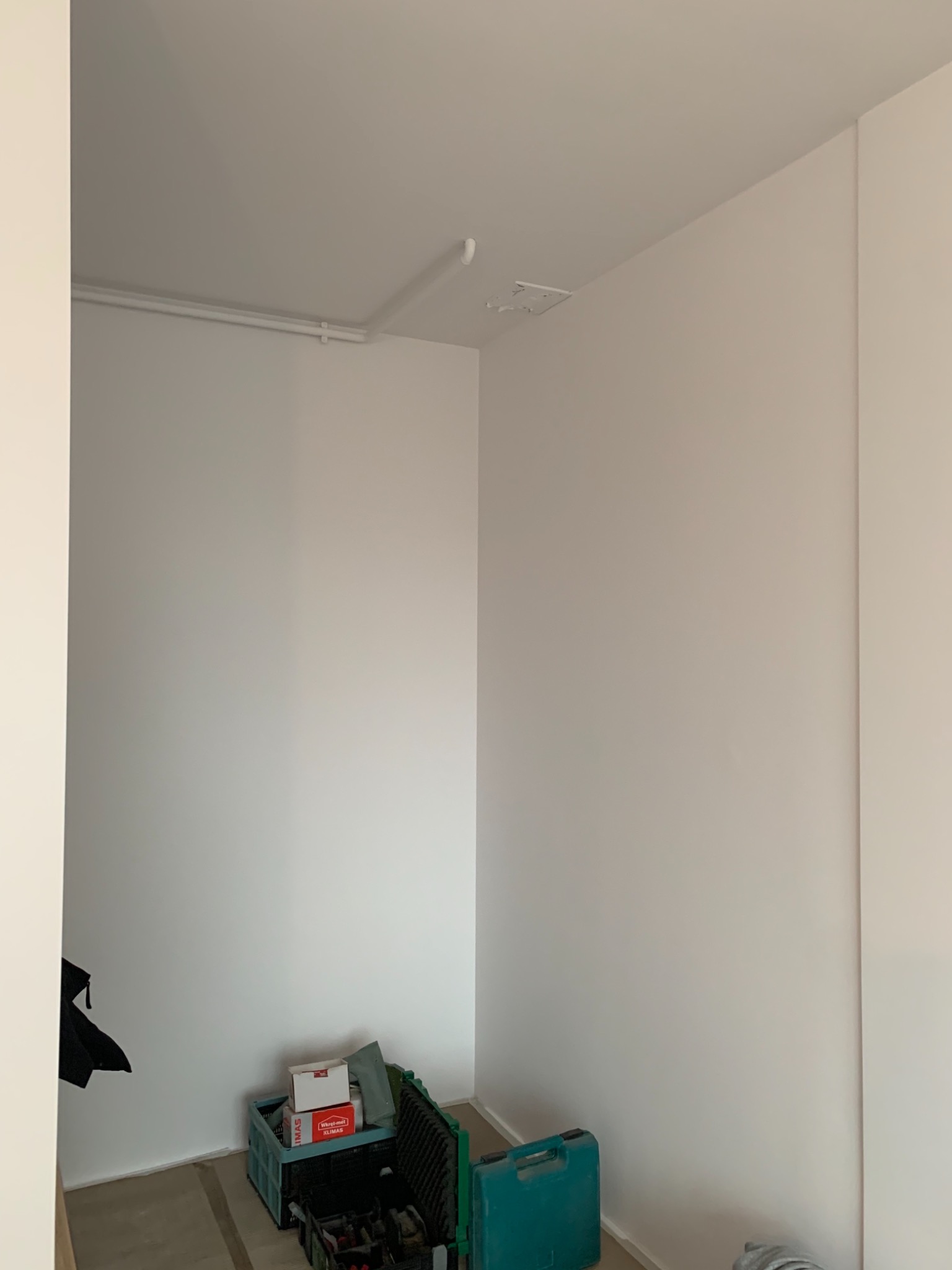 Wnętrze pokoju po świeżym malowaniu na biało, widoczne narzędzia i materiały malarskie w rogu pomieszczenia, rury pod sufitem.