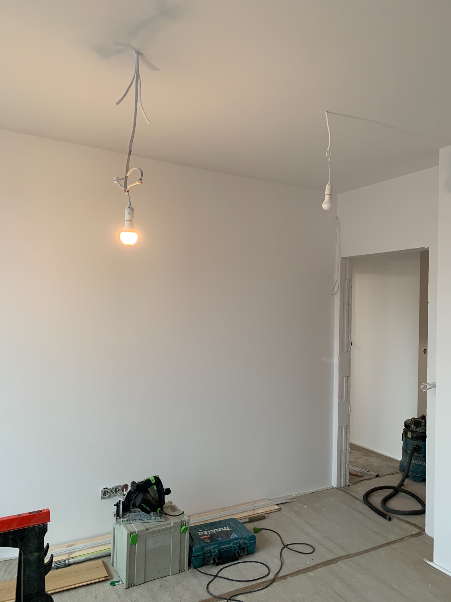 Wnętrze w trakcie remontu: świeżo pomalowana biała ściana, wiszące żarówki na kablach, narzędzia i skrzynki narzędziowe na podłodze, widoczny odkurzacz przemysłowy w przejściu do kolejnego...