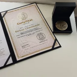 Certyfikat ukończenia studiów osteopatycznych w czarnej ramce, obok czarne pudełko z pamiątkową monetą Akademii Osteopatii.