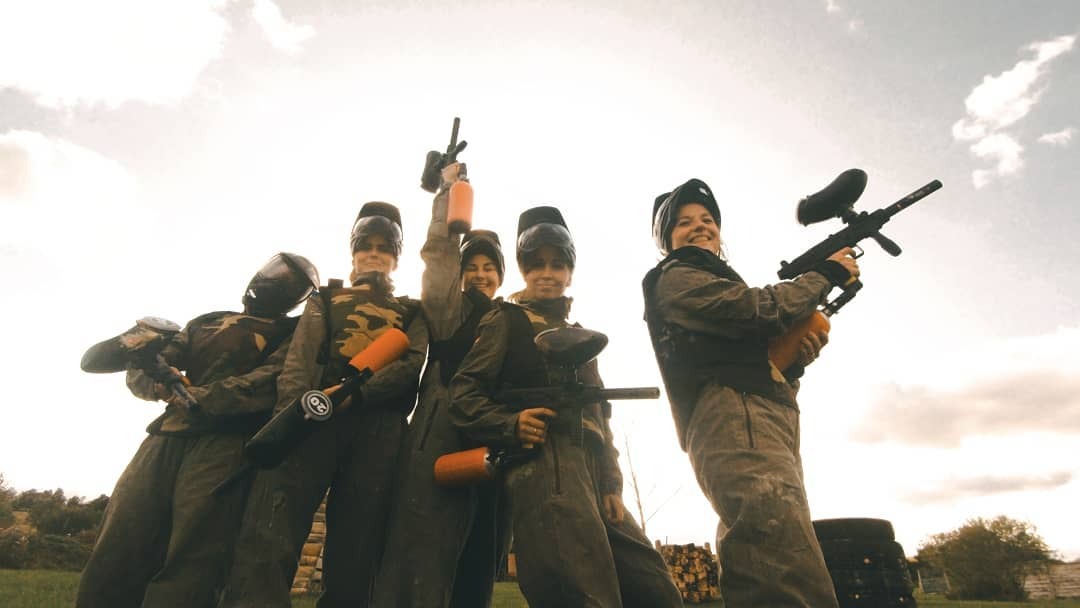 Grupa pięciu uśmiechniętych osób w strojach do paintballa, trzymających markery na tle trawiastego terenu i pochmurnego nieba.