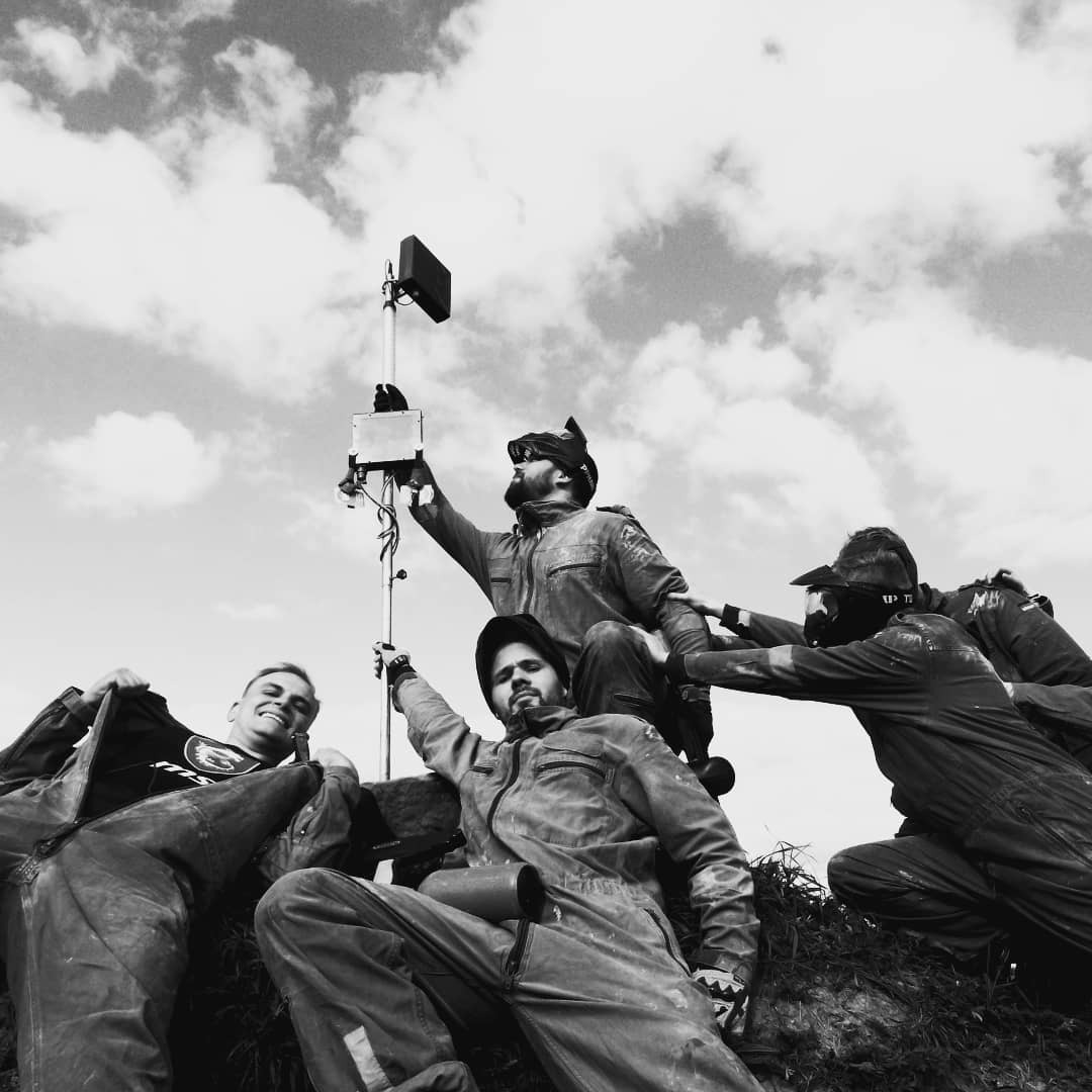 Czarno-białe zdjęcie grupy pięciu mężczyzn w kombinezonach po paintballu, pozujących na tle pochmurnego nieba, z jednym trzymającym antenę.