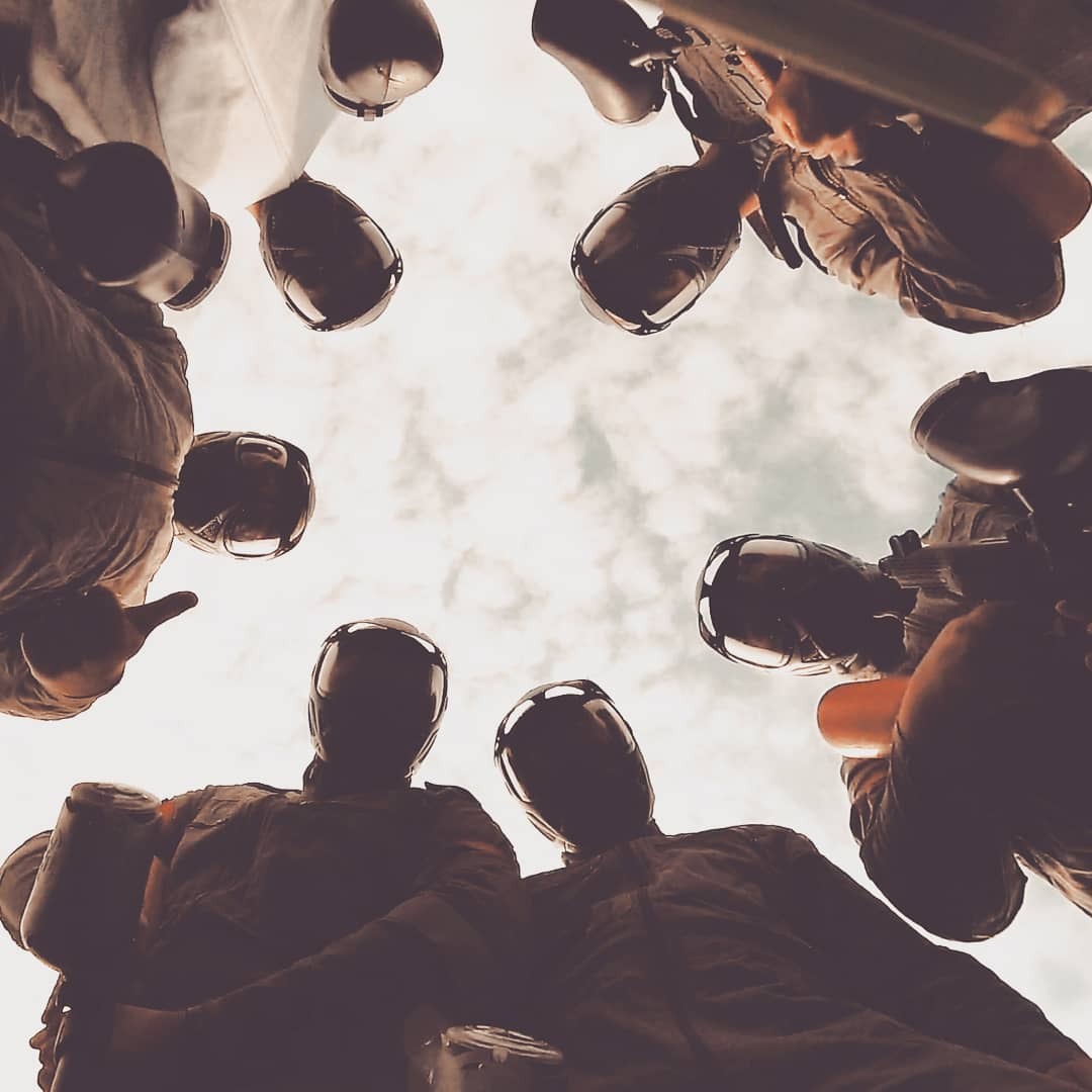 Grupa osób w kombinezonach i hełmach ochronnych, trzymających markery do paintballa, patrzy w dół z perspektywy żabiej, na tle pochmurnego nieba.