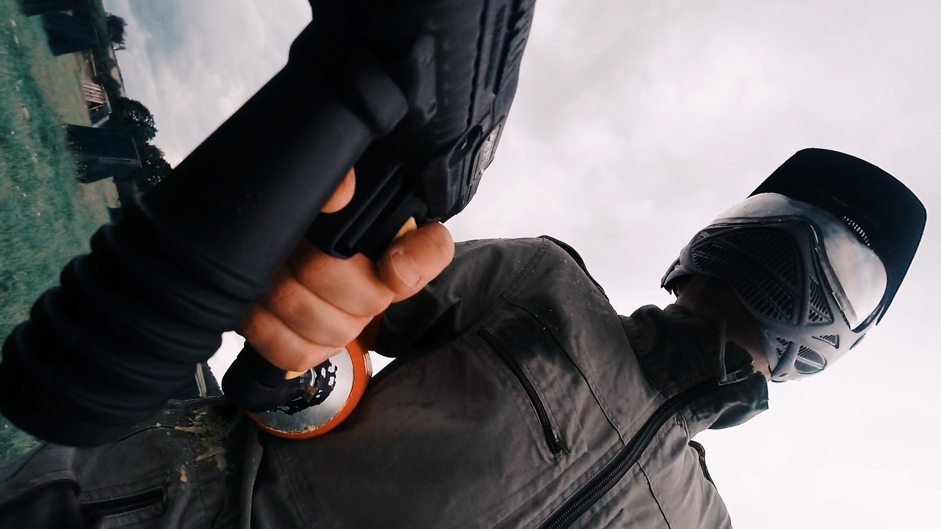 Gracz w paintball trafiony kulką, widok z perspektywy osoby strzelającej; w kadrze pistolet, pomarańczowa kulka i maska ochronna.