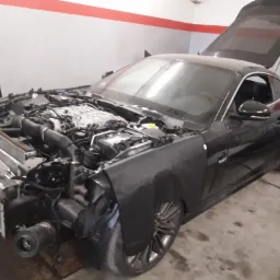 Czarny sportowy samochód z otwartą maską i bagażnikiem, widoczne elementy silnika, uszkodzony przód pojazdu, w tle ściana warsztatu.