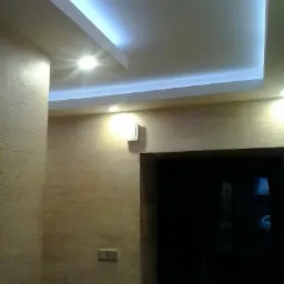 Beżowa ściana z fakturą, podświetlany sufit podwieszany z oświetleniem LED i wbudowanym punktowym światłem, widoczny włącznik światła i fragment ościeżnicy drzwi.