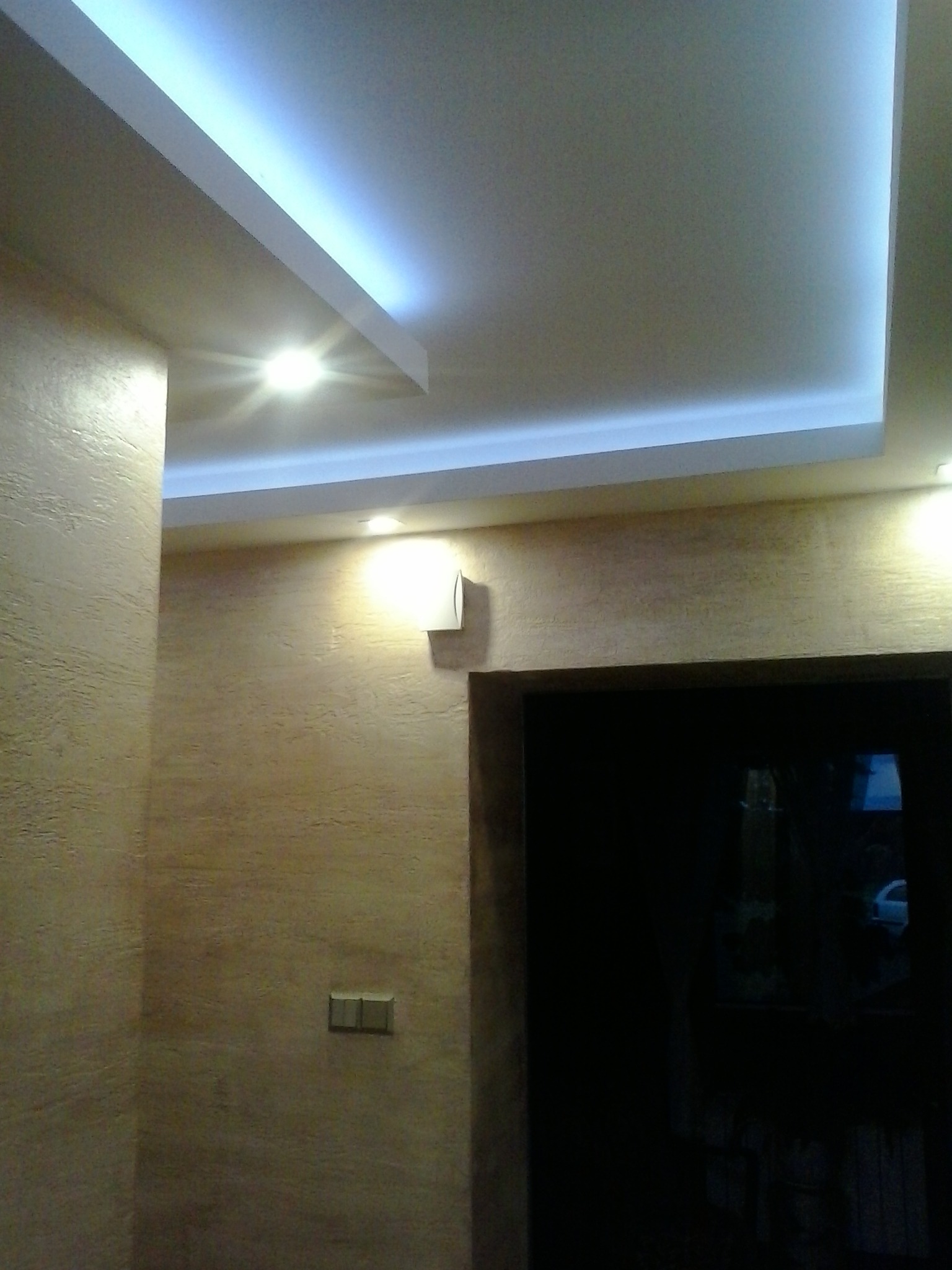 Beżowa ściana z fakturą, podświetlany sufit podwieszany z oświetleniem LED i wbudowanym punktowym światłem, widoczny włącznik światła i fragment ościeżnicy drzwi.