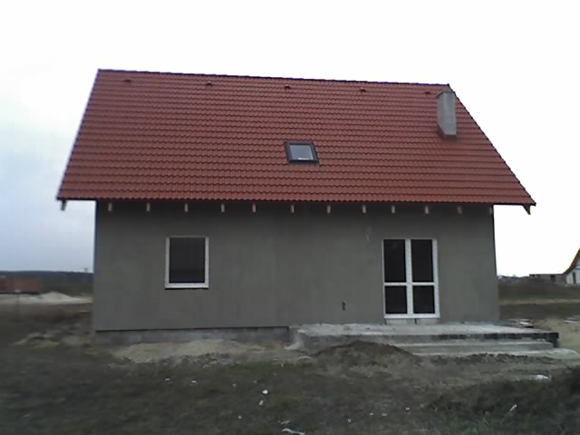 Surowy, nieotynkowany dom jednorodzinny z czerwoną dachówką i widocznym kominem, w trakcie budowy, z widocznymi fundamentami i schodami wejściowymi.