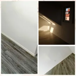 Pokój po remoncie z nowymi panelami podłogowymi imitującymi drewno, świeżo pomalowanymi białymi ścianami i listwami przypodłogowymi, widok z okna na sąsiedni budynek z czerwoną dachówką.