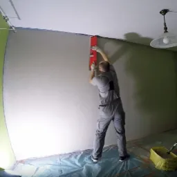 Mężczyzna w szarym kombinezonie wygładza ścianę czerwoną łatą, widoczna lampa sufitowa i zielona ściana w tle, podłoga zabezpieczona folią.