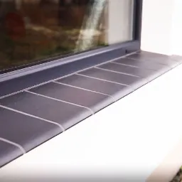 Zewnętrzny parapet okienny wyłożony ciemnoszarą płytką ceramiczną z jasnymi fugami, zamontowany na białej elewacji budynku.