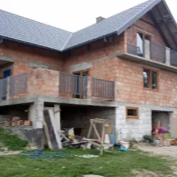 Surowy stan domu z czerwonej cegły w Nowym Sączu, z widocznymi balkonami i świeżo położonym szarym dachem, otoczony zielenią w trakcie prac wykończeniowych.