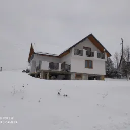 Niedokończony, dwupiętrowy dom z białymi ścianami i drewnianymi elementami, częściowo zasypany śniegiem w zimowy dzień.