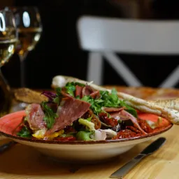 Apetyczna sałatka z szynką parmeńską, suszonymi pomidorami i świeżymi ziołami w ceramicznej misce z pomarańczowym brzegiem, podana z grissini i kieliszkami białego wina na drewnianym stole.