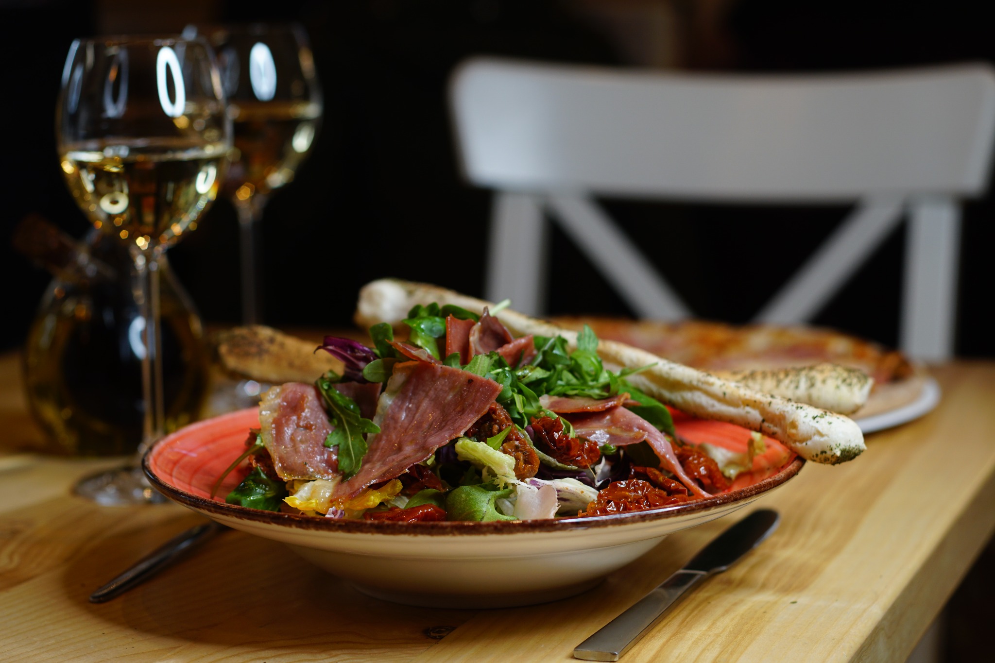 Apetyczna sałatka z szynką parmeńską, suszonymi pomidorami i świeżymi ziołami w ceramicznej misce z pomarańczowym brzegiem, podana z grissini i kieliszkami białego wina na drewnianym stole.