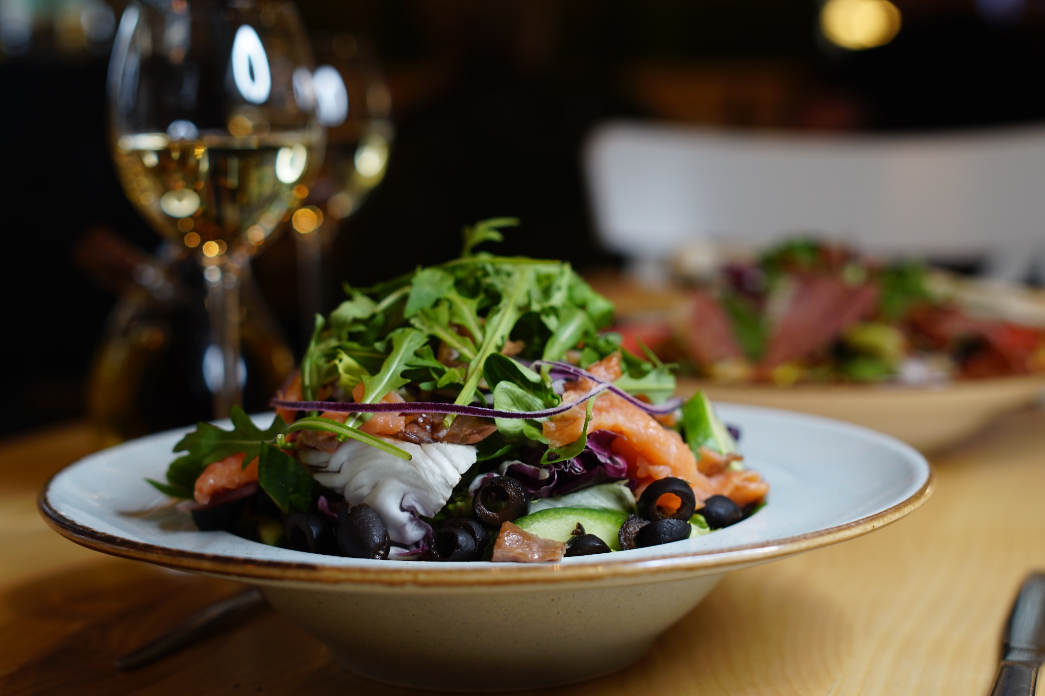 Apetyczna sałatka z łososiem, czarnymi oliwkami, rukolą i czerwoną cebulą, podana na białym talerzu z brązowym rantem, w tle dwa kieliszki białego wina i półmisek z wędlinami.